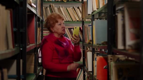 En äldre kvinna fotograferar med böcker i biblioteket. Selfie för sociala nätverk — Stockvideo