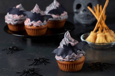 Cupcakes with colorful cream decorated with mastic bats and witch's brooms made of cheese and straw for Halloween party, dark background clipart