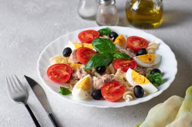 Makarnalı konserve ton balığı salatası, vişneli domates, siyah zeytin, kaynamış yumurta ve gri arka planda fesleğen..