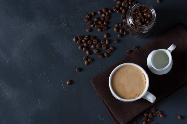 Beyaz bardakta sütlü kahve, koyu gri masada dağınık kahve çekirdekleri, kopyalama alanı, üst görünüm.