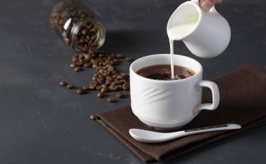 Süt koyu gri masada bir fincan kahveye dökülüyor, arka planda kahve çekirdekli bir kavanoz.