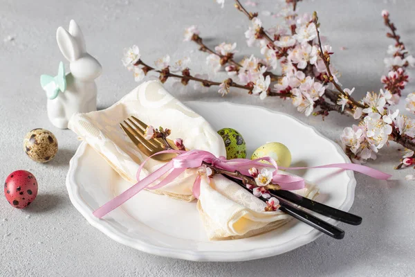 Puesta de mesa de Pascua con una rama de un árbol con flores, huevos de codorniz, una servilleta y cubiertos en un plato blanco en una mesa gris claro — Foto de Stock