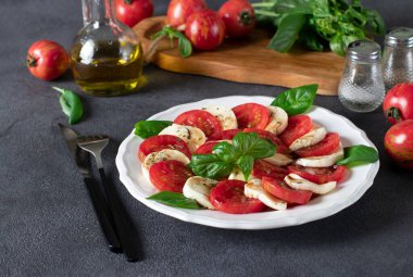 Dilimlenmiş domatesli İtalyan Caprese salatası, mozzarella peyniri, fesleğen ve gri arka planda beyaz tabakta zeytinyağı.