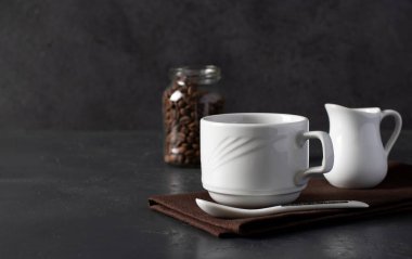 Beyaz bardakta sütlü kahve, koyu gri masada kavanozda kahve çekirdekleri...