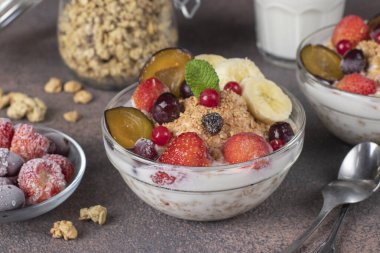 Doğal yoğurtlu gevrek müsli, donmuş böğürtlen, meyve ve fındıklar kahverengi arka planda cam kaselerde, sağlıklı kahvaltı.