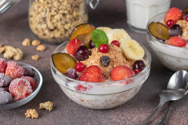 Granola chrupiące musli z jogurtem naturalnym, mrożone jagody i owoce i orzechy w szklanych miskach na brązowym tle, zdrowe śniadanie — Zdjęcie stockowe