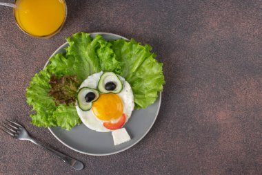Çocuklar için eğlenceli yemek. Yağda yumurta, salatalık, domates, siyah zeytin ve gri tabakta salata yapraklarından yapılmış gülen surat. Çocuklar için yaratıcı, sağlıklı kahvaltı. Boşluğu kopyala.