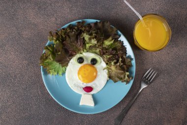 Çocuklar için eğlenceli yemek. Kahverengi arka planda kızarmış yumurta, salatalık, turp ve marul yapraklarından yapılmış gülen surat. Çocuklar için yaratıcı sağlıklı kahvaltı
