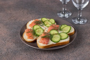 Kahverengi zemin üzerinde tuzlanmış somon, salatalık ve dereotlu sandviçler.