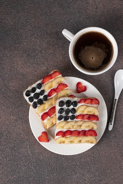 American flag sandwiches with strawberries, banana and cream cheese on rye bread and cup of coffee on brown background. Independence Day breakfast idea, Vertical format