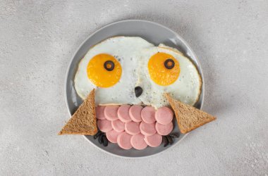 Çocuklar için eğlenceli yiyecekler. Sosisli sahanda yumurtadan çıkan komik baykuş, gri arka planda ekmek ve siyah zeytin. Yaratıcı, sağlıklı kahvaltı, Top view