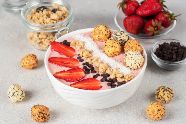 Smoothie bowl with strawberries, granola, chocolate, coconut flakes and crunchy honey balls in white bowl on light gray background