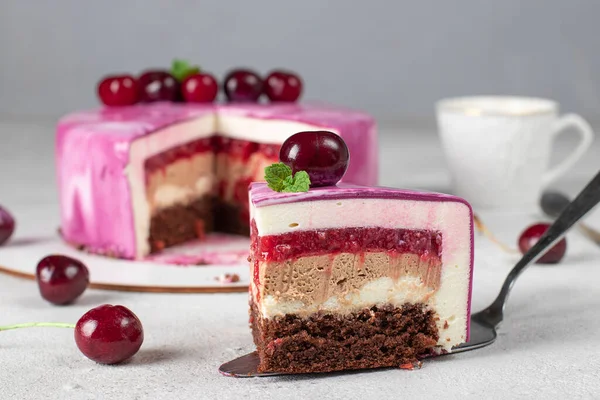 Piece of mousse cake with chocolate biscuit, vanilla and cream mousse, cherry confit and milk chocolate ganache on gray background