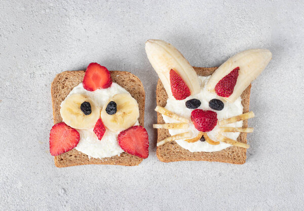 Two sweet toasts for kids in shape of chick and rabbit with strawberries, banana, cream cheese and coconut flakes on gray background, Top view