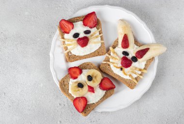 Three sweet toasts for kids in shape of cat, rabbit and chick with strawberries, banana, cream cheese and coconut flakes on white plate on gray background