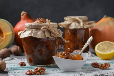 Homemade pumpkin jam with lemon and walnuts in jars and bowl on gray background