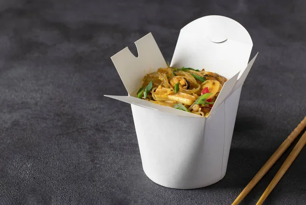Noodles with seafood and vegetables in takeaway box on dark gray table