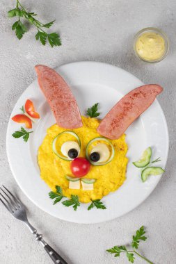 Fun food for kids breakfast - cute hare made from scrambled eggs, sausage, cheese, tomato, cucumber and black olives, Top view, Vertical format