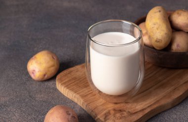 Vegan potato milk in glass and potato in plate on wooden board on brown background