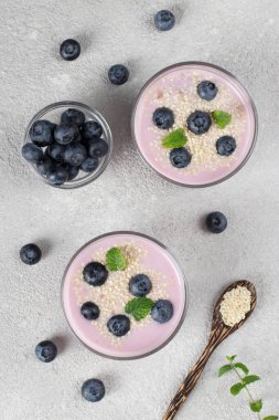 Thick yogurt with blueberries and sesame seeds in glasses, as well as in wooden spoon on llight gray background, Top view