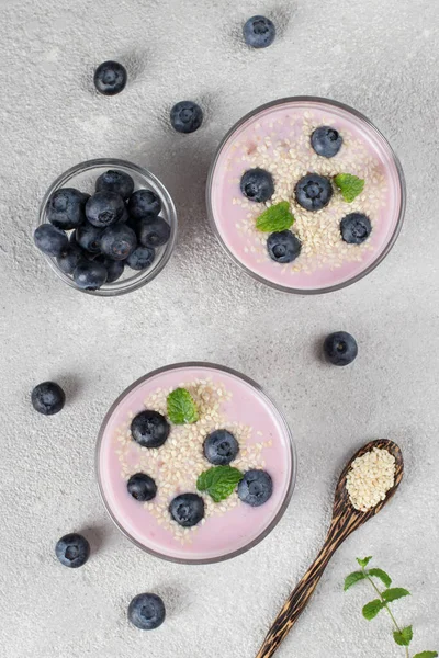 Thick yogurt with blueberries and sesame seeds in glasses, as well as in wooden spoon on llight gray background, Top view