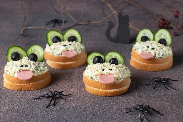 Sandwiches with cucumber, sausage, black olives, egg and melted cheese in the shape of frogs on brown background - creative idea for Halloween appetizer