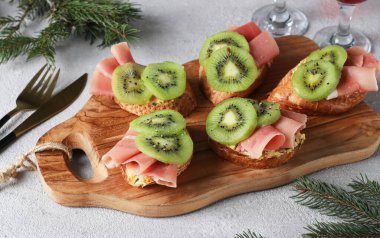 Sandwiches with soft cheese, jamon and kiwi on wooden board on light gray background