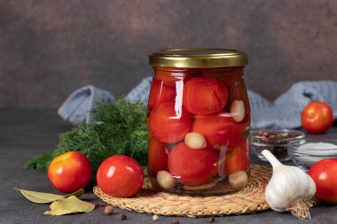 Pickled cherry tomatoes with garlic in glass jar on brown background