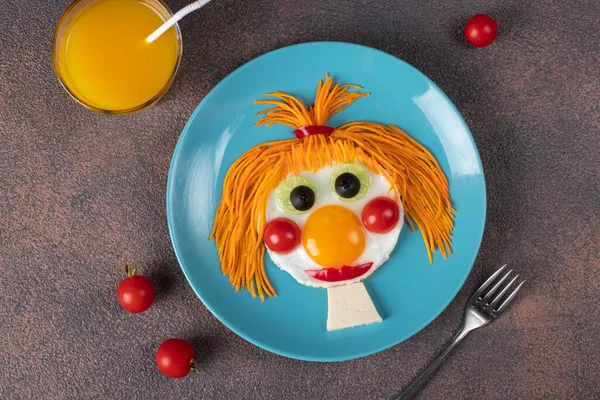 Fun Food for kids - smiling face ginger girl made from fried eggs, carrot, cucumbers and cherry tomatoes on blue plate. Creative healthy breakfast for children