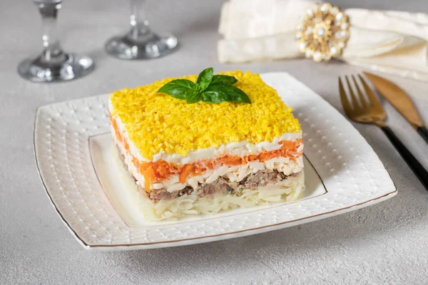 Traditional Russian puff salad Mimosa with canned fish, potatoes, cheese, carrots and eggs on light gray background