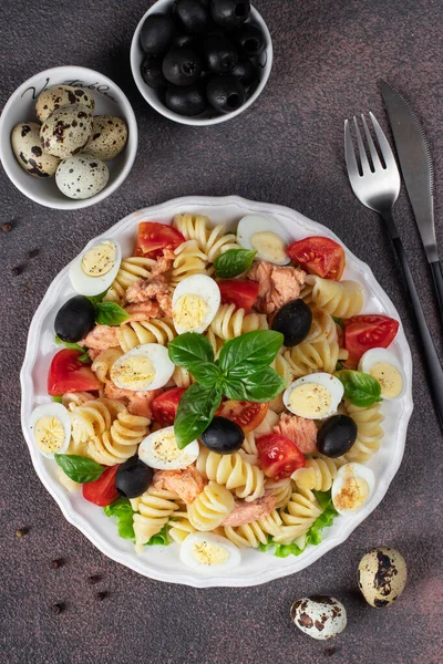 Canned salmon salad with pasta, cherry tomatoes, quail eggs, basil and black olives on brown background, Top view