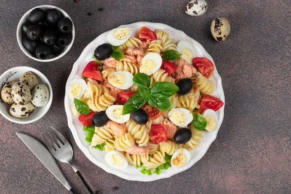 Canned salmon salad with pasta, cherry tomatoes, quail eggs, basil and black olives on brown background, Top view