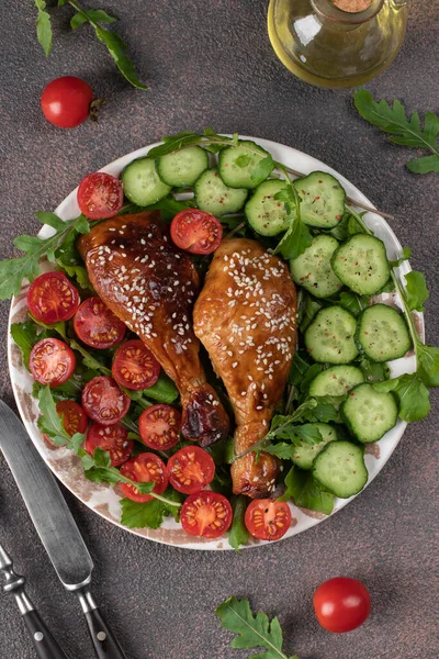 Baked chicken drumsticks with cherry tomato, cucumber and arugula salad on brown background, Top view