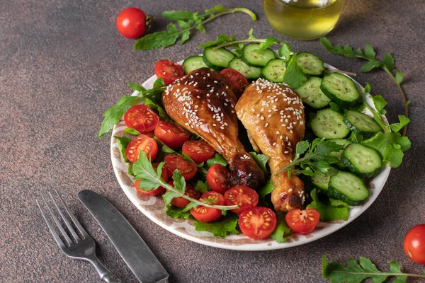 Baked chicken drumsticks with cherry tomato, cucumber and arugula salad on brown background