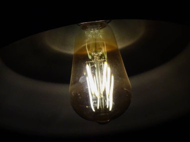 Geleneksel bir ampul yakın plan ve bir kat cam ve loş ışık. Lamba çatıda asılı. Karanlıkta parlıyor.