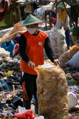 Ponorogo, Endonezya - 10 08 2021: Turuncu gömlekli ve yeşil şapkalı bir adam bir çuval dolusu plastik atığı elinde tutuyor..