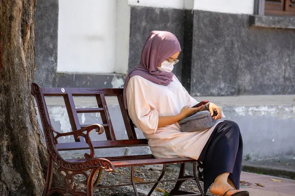 Surabaya, Jawa Timur / Endonezya - 09 28 2020: Kovid-19 salgını sırasında maske takan Müslüman kadınlar sadece sağlık protokollerini uygulamak için banklarda oturuyorlar. Kadın cep telefonuna bakıyor..