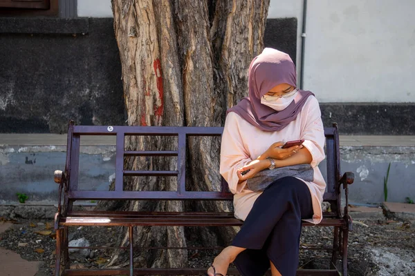 スラバヤ ジャワ ティムール インドネシア 2020年9月28日 マスクをしたイスラム教徒女性がベンチに座って 流行期に健康プロトコルを実装しています 女性は携帯電話を見て — ストック写真