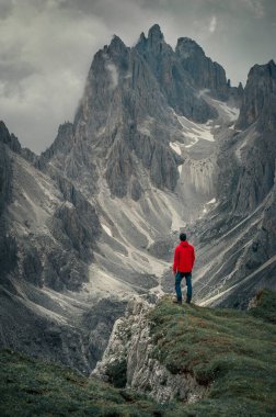Man with red jacket in front of mountain peaks in the Dolomite Alps in South Tyrol with dramatic cloudy sky, Three Peaks Nature Reserve, Italy clipart