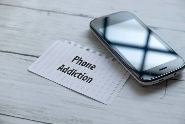 Selective focus of mobile phone and paper written with Phone Addiction on white wooden background.