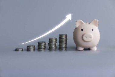 A stack of ascending coins with piggy bank and glowing white arrow on grey background.