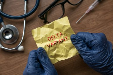 A hand wearing blue glove opening a crumpled yellow memo note written with Delta Variant.