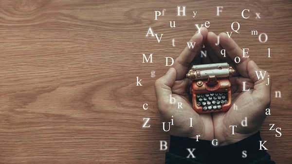 A hand holding toy typewriter with alphabet flying around on wooden table with copy space.