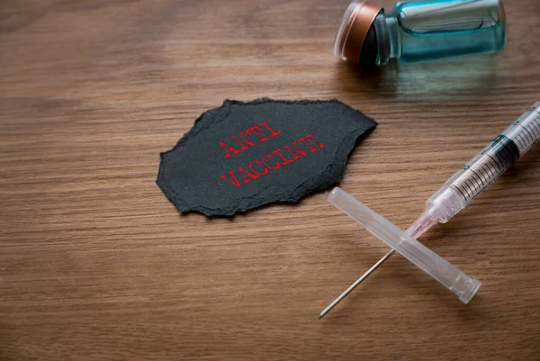 A bottle of vaccine, syringe and a piece o black paper written with Anti Vaccine on wooden background.