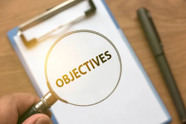 A hand magnifying a words of Objectives from a white paper using magnifying glass.