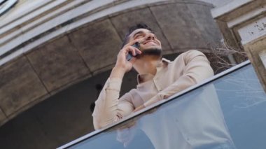 Dipteki manzara mutlu mutlu gülümseyen başarılı kullanıcı ispanyol Arap adam teras ofis merkezinde cep telefonuyla konuşuyor. Şehir merkezindeki modern şirkette balkonda akıllı telefon konuşması.