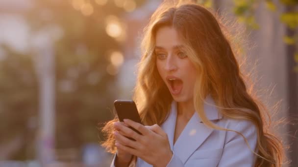 Glad kaukasiska affärskvinna njuta av framgång ser i mobiltelefon i stadens sol bakgrund. Närbild glad flicka läser goda nyheter i smartphone gör ja hand gest. Lady firar online seger — Stockvideo