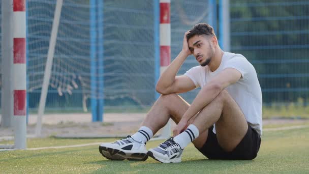 Sfrustrowany zdenerwowany zamyślony bliskowschodni piłkarz siedzący na trawie boiska do piłki nożnej przed bramką, rozważający koniec gry, tysiącletni sportowiec w letniej odzieży sportowej niezadowolony wynik, rozczarowane wyniki — Wideo stockowe