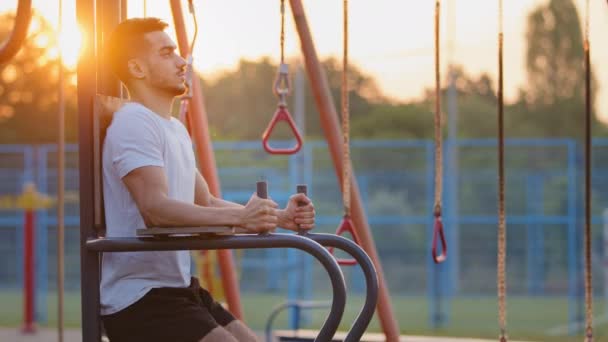 Ortadoğulu atletik genç vücut geliştirici. Açık şehir spor sahasında çalışırken karın kaslarını çalıştırıyor. Yetişkin, yakışıklı, güçlü, sportif Hintli adam aletleri kullanarak kaslarını şişiriyor. — Stok video