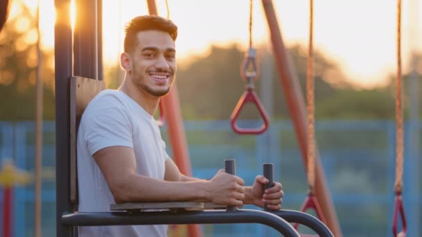 Портрет привлекательный тысячелетний ближневосточный арабский спортивный парень делает упражнения на открытом воздухе на спортивной площадке. Молодой взрослый индийский спортсмен улыбается приветливо стоя рядом с оборудованием во время утренней тренировки — стоковое видео